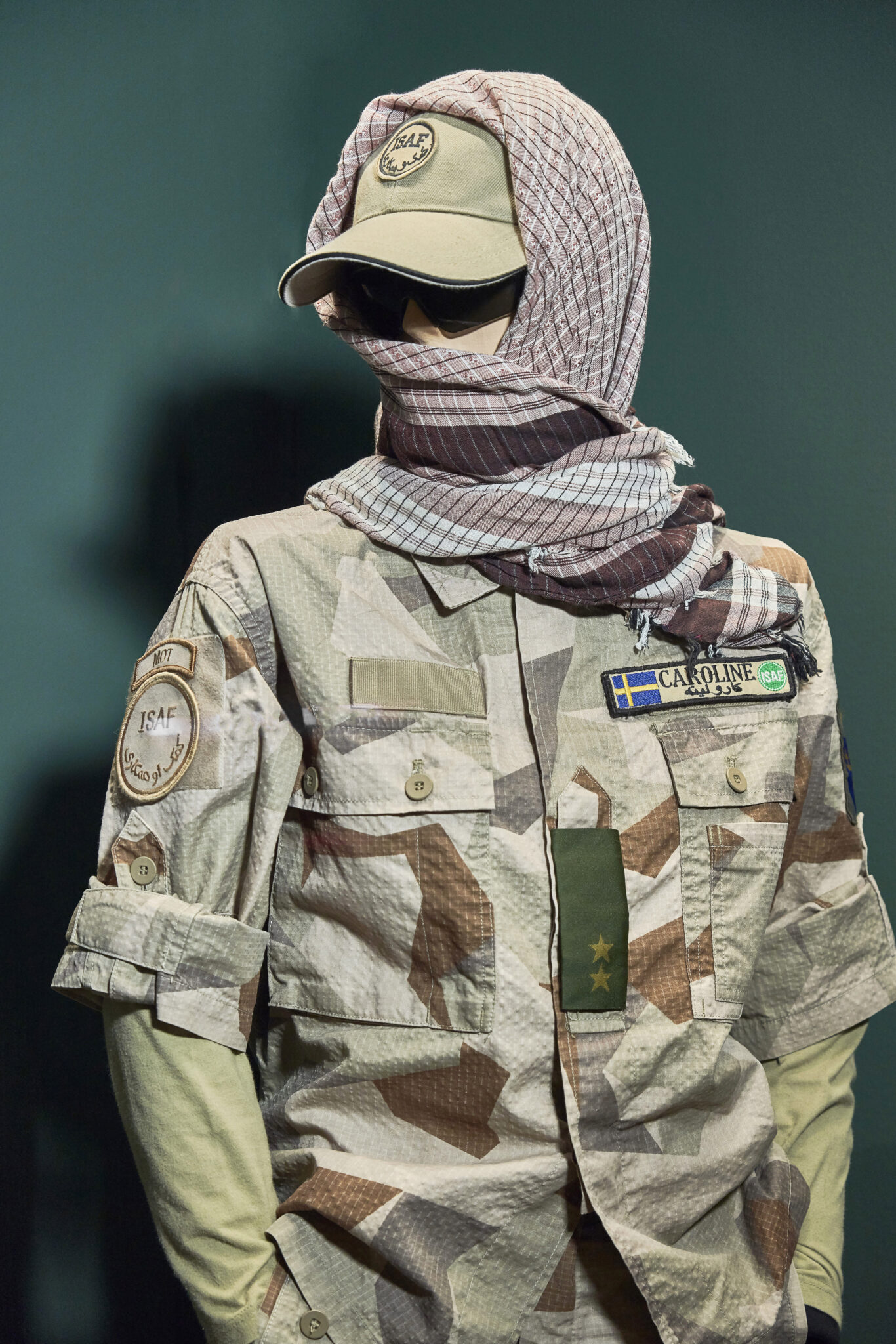 En docka i verklig storlek kl&auml;dd i kamouflagef&auml;rgad uniform, gr&ouml;n keps och en sjal i jordn&auml;ra f&auml;rger draperad om huvudet.