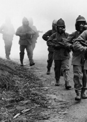Soldater går på led med vapen i famnen och gasmask över ansiktena.