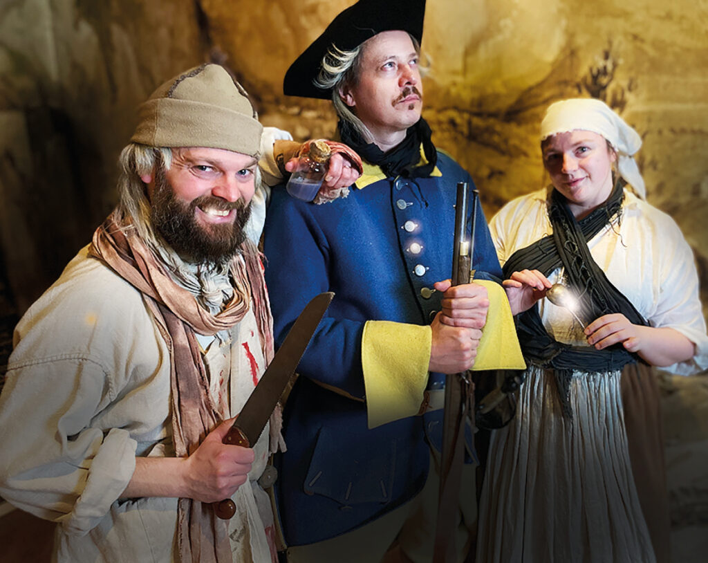 Tre karaktärer i 1700-tals kläder. Varav en smutsig fältskär som ler lurigt, en stolt soldat med blanka knappar och en trasig marketenterska med en blank sked.