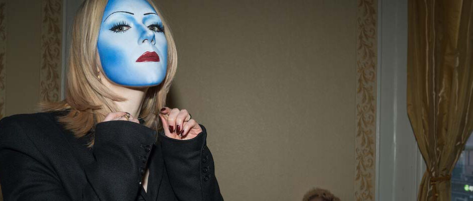 En kvinna med ansiktet helt m&aring;lat i ljusbl&aring;tt, som en mask.